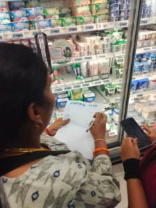 An over-the-shoulder photo of a workshop participant inside a supermarket aisle. She is holding a pen and a shopping list, writing on it as she looks at dairy products in a refrigerated section. The hand of another person holding a smartphone is visible next to her.