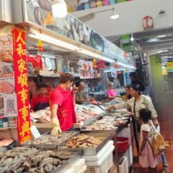 A vibrant photo from inside a bustling wet market. A female workshop participant and a young girl stand at a fresh seafood stall, interacting with the vendor. The stall is filled with fresh fish and prawns on ice. Other people are visible in the background, observing the interaction.