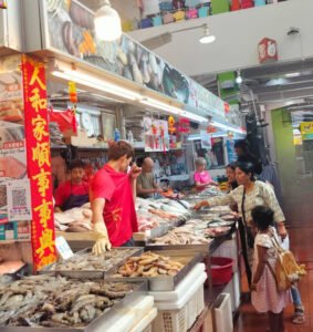A vibrant photo from inside a bustling wet market. A female workshop participant and a young girl stand at a fresh seafood stall, interacting with the vendor. The stall is filled with fresh fish and prawns on ice. Other people are visible in the background, observing the interaction.