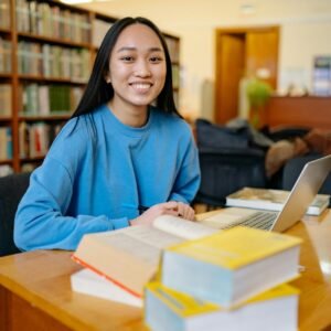 A young woman sits at a desk in a library, smiling at the camera with an open book and a laptop in front of her.