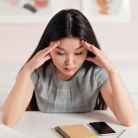 A young woman with a worried expression holds her head in her hands while looking down at a notebook and phone.