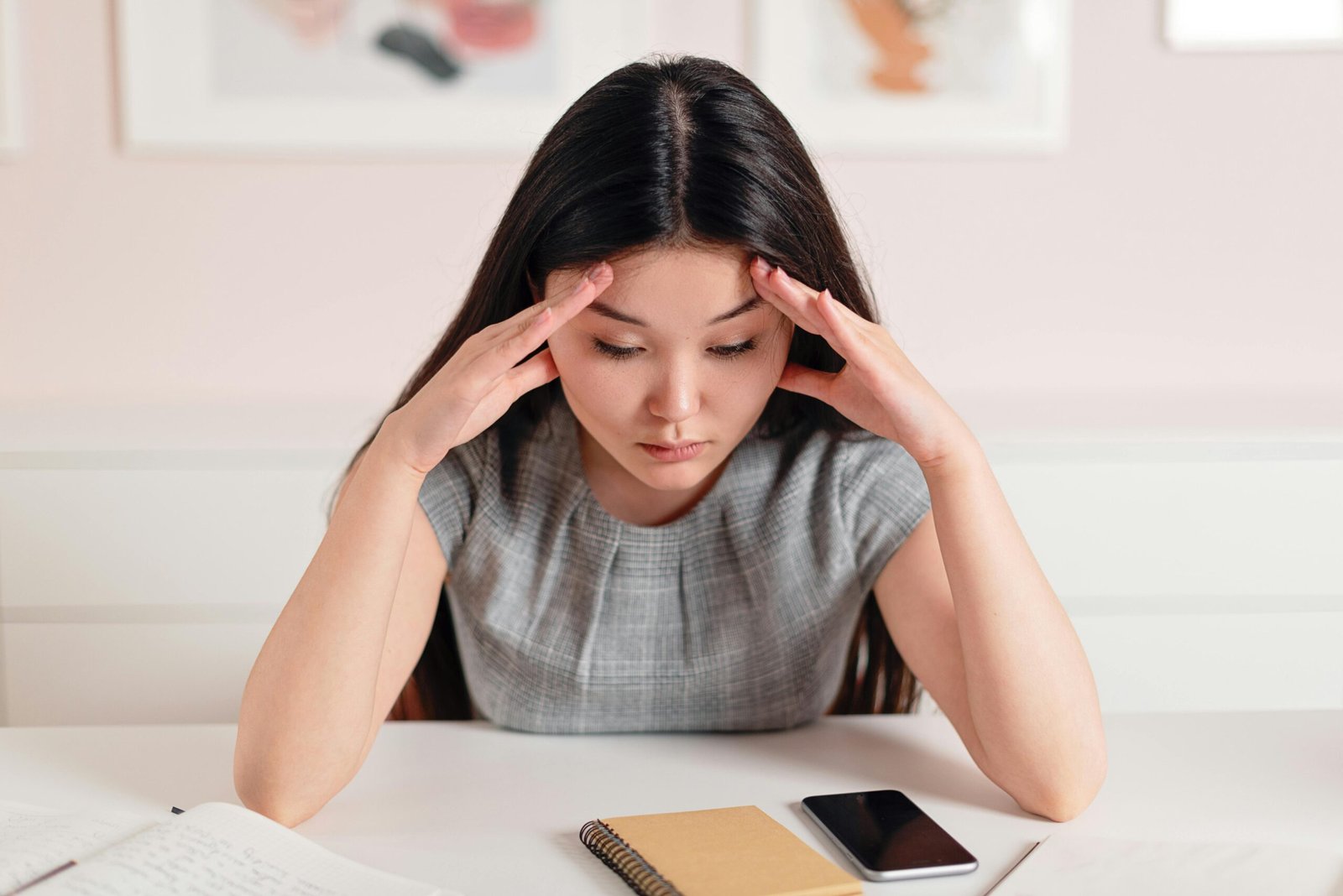 A young woman with a worried expression holds her head in her hands while looking down at a notebook and phone.