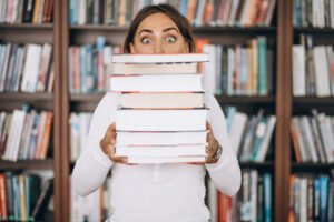 A woman with a surprised expression holds a tall stack of books, hiding her face, in front of a bookshelf.