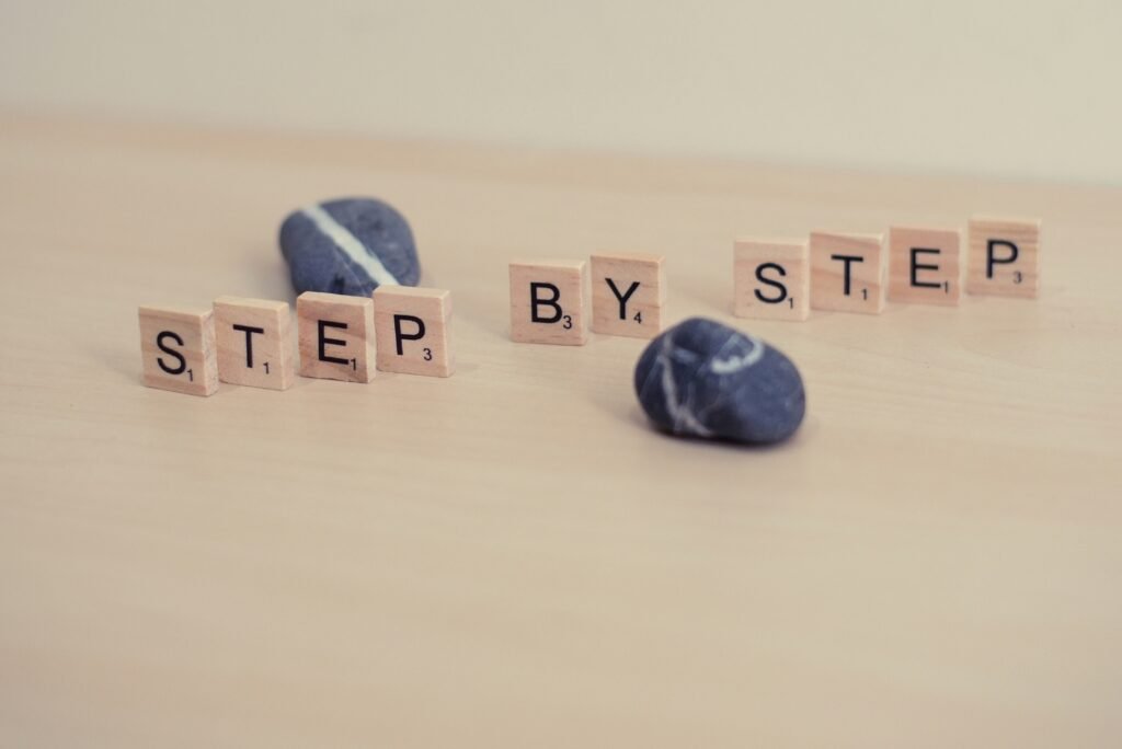 Wooden Scrabble tiles spelling "STEP BY STEP" on a light wood surface, interspersed with smooth grey stones
