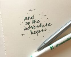 A close-up photo of a notebook with the handwritten phrase "and so the adventure begins" in black and green ink, next to a silver fine-tip pen and a white marker.