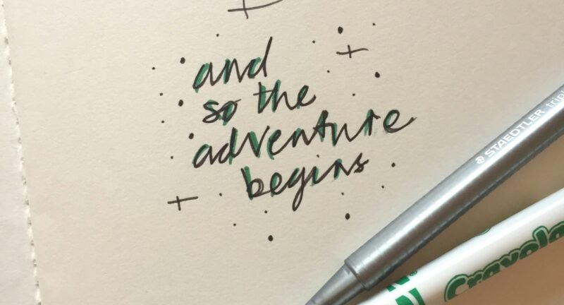 A close-up photo of a notebook with the handwritten phrase "and so the adventure begins" in black and green ink, next to a silver fine-tip pen and a white marker.