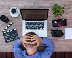 Top-down view of a person sitting at a wooden desk with a laptop, camera, and film clapperboard, holding their head in frustration.