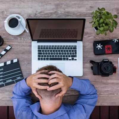 Top-down view of a person sitting at a wooden desk with a laptop, camera, and film clapperboard, holding their head in frustration.