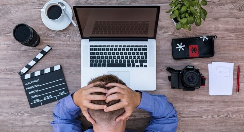 Top-down view of a person sitting at a wooden desk with a laptop, camera, and film clapperboard, holding their head in frustration.