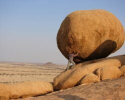 Financial Resilience - How To Build A Foundation That Lasts A person in a desert landscape putting their weight against a massive, round boulder on a slope, symbolizing the strength and effort involved in building financial resilience.