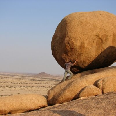 A person in a desert landscape putting their weight against a massive, round boulder on a slope, symbolizing the strength and effort involved in building financial resilience.