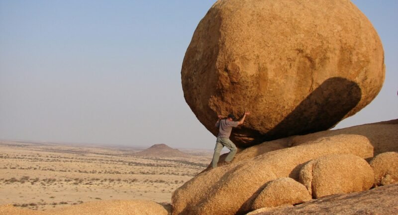 A person in a desert landscape putting their weight against a massive, round boulder on a slope, symbolizing the strength and effort involved in building financial resilience.