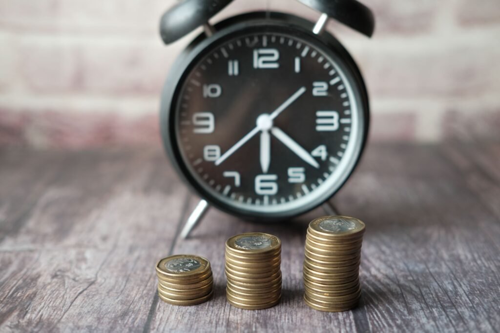 Three growing stacks of coins in front of a blurred vintage alarm clock, illustrating the concept of money versus time.