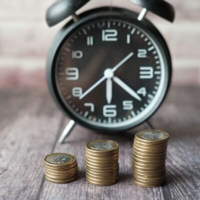 Three growing stacks of coins in front of a blurred vintage alarm clock, illustrating the concept of money versus time.