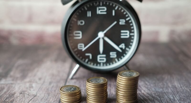 Three growing stacks of coins in front of a blurred vintage alarm clock, illustrating the concept of money versus time.